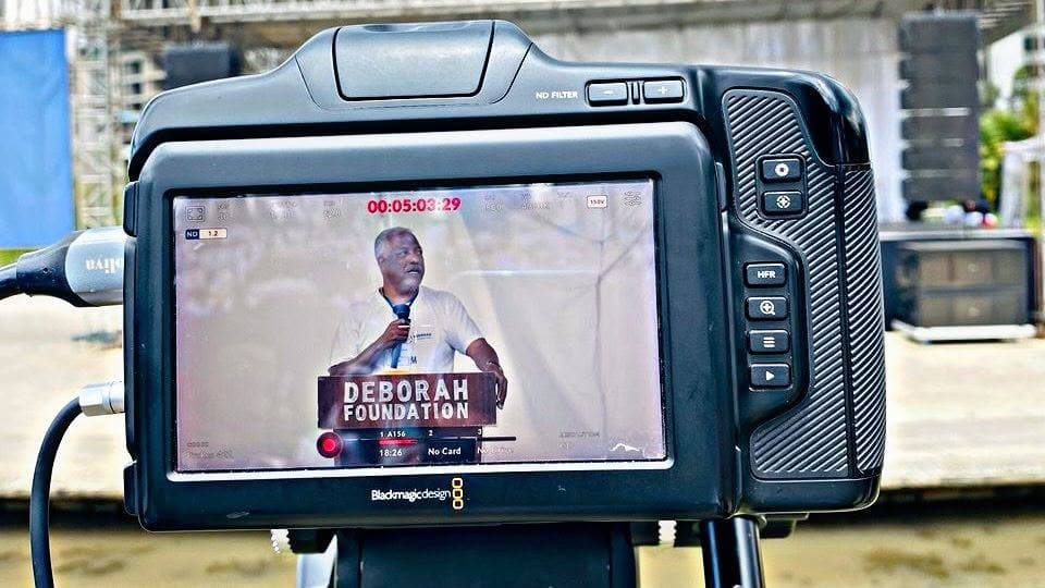 Camera monitor showing a titled live feed of a speaker at an outdoor stage event